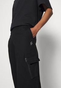 Pantaloni cargo neri realizzati in tessuto liscio, con una tasca con zip sul lato e un logo sul davanti. Design semplice e funzionale.