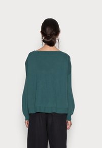 Tiefer Pullover in Türkis mit weitem Ausschnitt, lässiger Passform, gerippten Bündchen und Saum. Gestrickte Textur, getragen mit schwarzen, schmal zulaufenden Hosen.