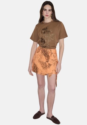 Mujer con top marrón de manga corta con bordado floral, falda cruzada naranja con estampado, pendientes colgantes y zapatos tipo slip-on marrón oscuro.