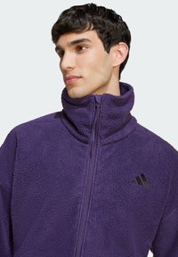 Giacca in pile viola con colletto alto, chiusura con zip e logo nero. Il tessuto testurizzato presenta un aspetto morbido e soffice.