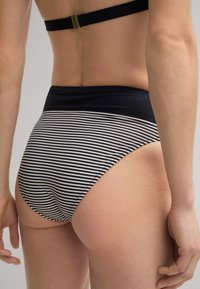 Vrouw in een zwart-wit gestreepte bikini met hoge taille, met een zwart bovenstuk, gezien van achteren tegen een effen achtergrond.