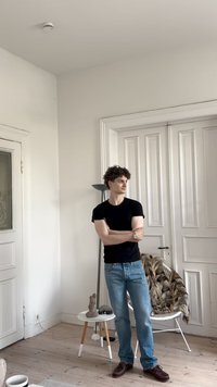 Fiatal férfi fekete pólóban és farmert viselve áll, karba tett kézzel egy minimalista szobában, fehér falakkal, fapadlóval és modern bútorokkal.