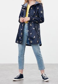 Marinblå regnjacka med hundmönster, långa ärmar och huva. Lätt, glänsande material. Bärs över en gul topp och kombineras med ljusa jeans och sneakers.