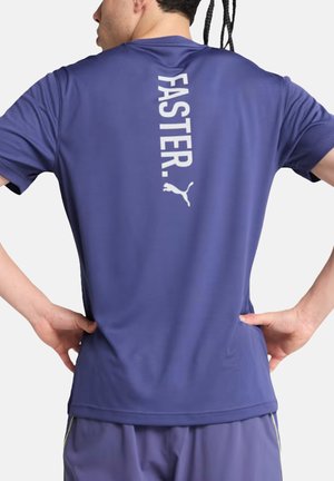 Uomo con maglietta sportiva blu e pantaloncini con la scritta "FASTER." e il logo Puma verticali sulla schiena, mani sui fianchi, di spalle alla fotocamera.