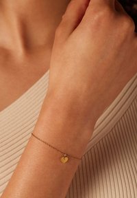 Gouden kettingarmband met een klein hartje, gedragen aan een hand, tegen een textuur van beige achtergrond. Eenvoudig en delicaat ontwerp.