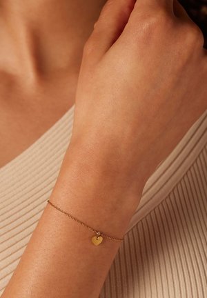 Gouden kettingarmband met een klein hartje, gedragen aan een hand, tegen een textuur van beige achtergrond. Eenvoudig en delicaat ontwerp.