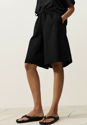 Personne portant un short ample noir jusqu'aux genoux avec une taille élastique et des tongs noires, debout sur un sol carrelé.