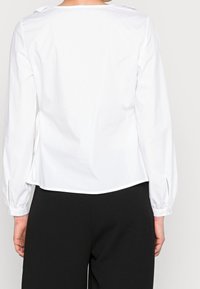 Blusa bianca con maniche lunghe, caratterizzata da un ampio scollo e polsini plissettati. Tessuto liscio con legature regolabili ai lati.