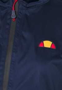 Marinblå tygjacka med framdragkedja, som har en liten, flerfärgad logotyp—gul, röd och orange—på den vänstra bröstområdet.