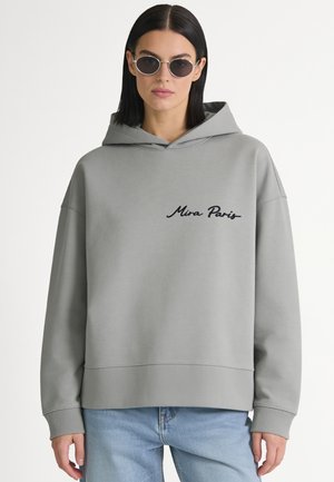 Vrouw met stijl donker haar draagt ronde zonnebril, een grijze hoodie met tekst "Mina Paris" en lichtblauwe spijkerbroek, kijkt recht vooruit op witte achtergrond.