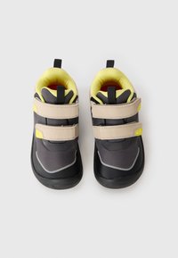 Chaussures de sport noires et grises avec des sangles Velcro beiges et des accents jaunes. Texture lisse avec une semelle en caoutchouc et un intérieur rembourré.