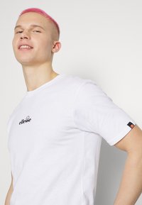 Weißes Baumwoll-T-Shirt mit einem schwarzen "ellesse"-Logo auf der Brust und einem Logo-Label am Ärmel. Kurze Ärmel, Rundhalsausschnitt.