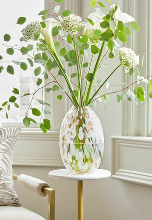 Vaso di vetro trasparente con composizione floreale bianca e verde su un tavolino rotondo bianco e oro vicino a una finestra.