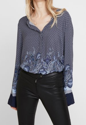 Marineblauwe blouse met een patroon en een bloemenprint aan de zoom, voorzien van een V-hals en lange mouwen. Gecombineerd met nauwsluitende zwarte leren broek.