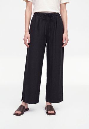 Persona con pantalones negros holgados con aberturas en los tobillos, una camiseta blanca y sandalias negras sin cordones de color marrón oscuro sobre un fondo blanco liso.
