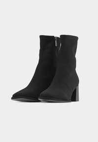 Ζευγάρι μαύρες μπότες μεσαίο υ μήκους από ελαστικό ύφασμα, με χοντρά τακούνια και φερμουάρ στο εσωτερικό μέρος, σε απλό λευκό φόντο.