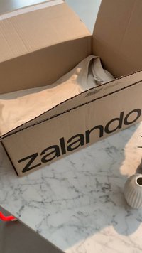 Caixa de cartão rotulada "zalando," parcialmente aberta, revelando um item de vestuário bege no interior, posicionada sobre uma bancada de mármore com um vaso próximo.