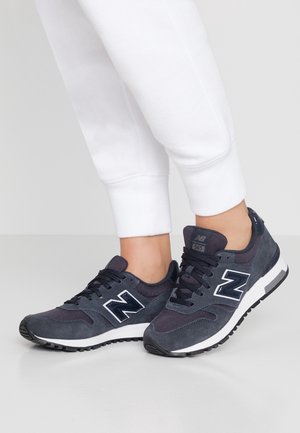 Osoba w ciemnoszarych sneakersach New Balance 565 z białymi podeszwami i białych spodniach dresowych na jednolitym szarym tle.