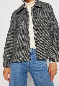 Kurtka w wzór houndstooth w czerni i bieli, z kołnierzykiem, długimi rękawami i pięcioma czarnymi guzikami z przodu.