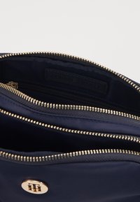 Custodia in tessuto blu navy con zip dorata, tasche interne e un bottone logo dorato. La fodera testurizzata offre contrasto e resistenza.