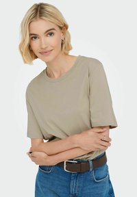 Beige bomulds-T-shirt med rund hals og korte ærmer, parret med blå denimjeans og et brunt bælte. Glat tekstur, afslappet design.