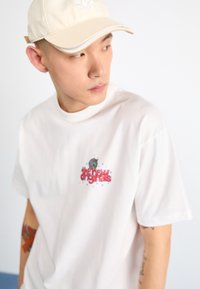 White short-sleeve t-shirt featuring a graphic design with red text "the new originals" and blue accents. Pairing with a beige baseball cap.