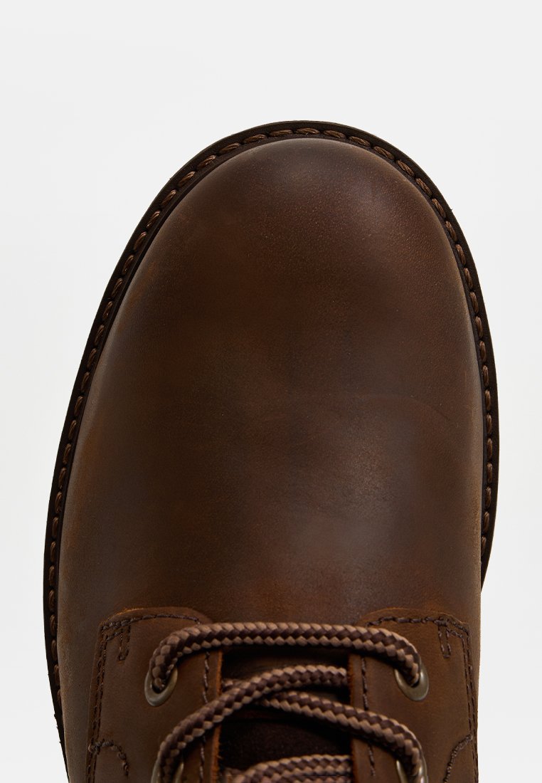 Brauner Lederstiefel-Zeh mit detaillierter Naht und geflochtenen Schnürsenkeln auf weißem Hintergrund.