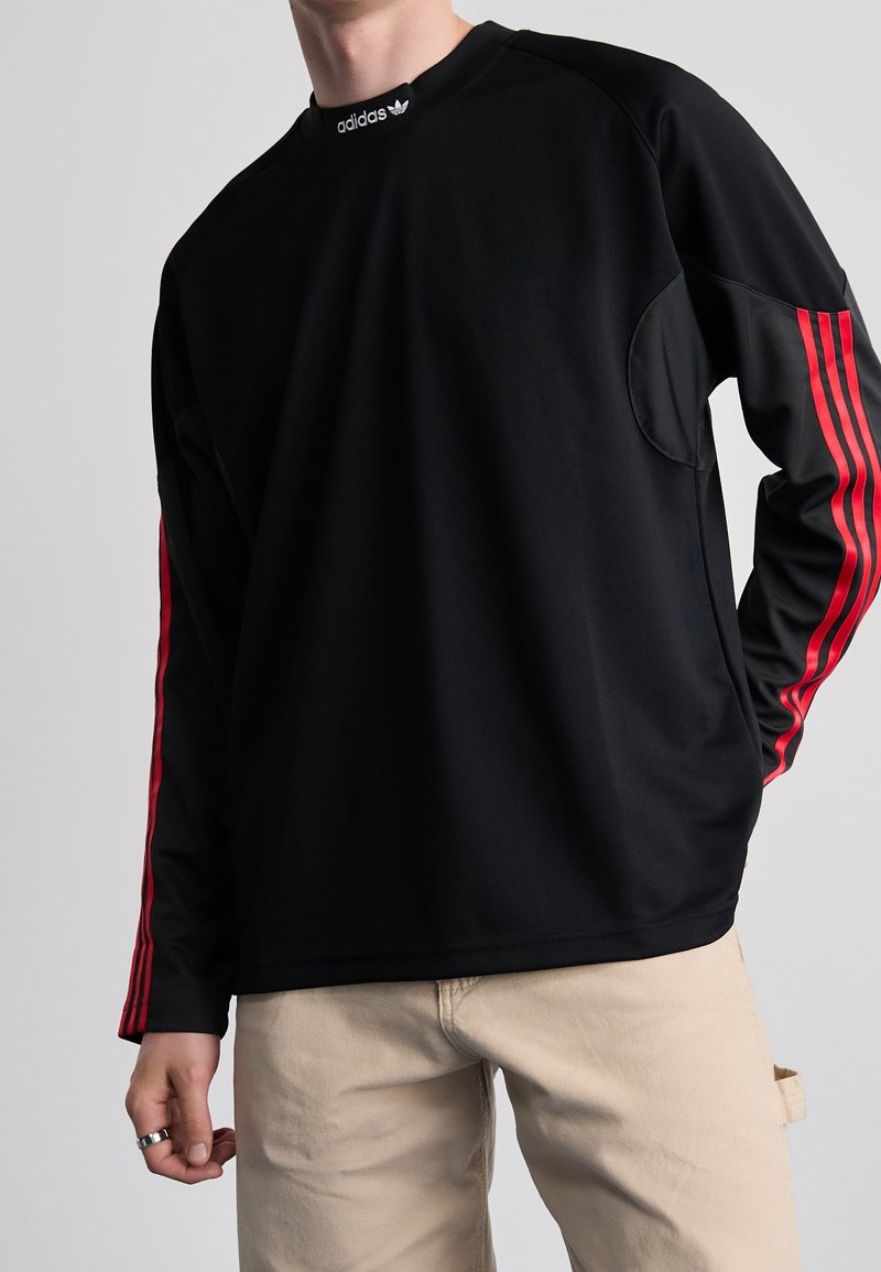 Person wearing black long-sleeve Adidas top with red stripes on sleeves and logo on collar, paired with beige pants.