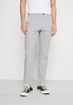 Man draagt slim-fit grijze en witte verticale gestreepte broek met zwarte knoop, wit T-shirt en groene lage sneakers.