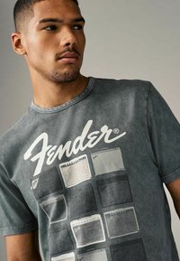 Gray cotton t-shirt featuring a large "Fender" logo and graphic of various guitar amplifiers in muted colors, short sleeves, and crew neck.