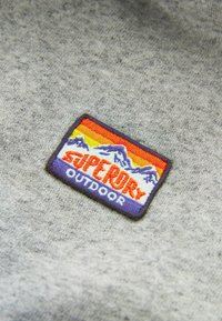 Grauer Sweatshirtstoff mit einem farbenfrohen, bestickten Aufnäher, der Berge, einen Sonnenuntergangsverlauf und den fett gedruckten Schriftzug „SUPERDRY OUTDOOR“ zeigt.