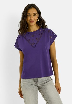 Mujer con cabello oscuro y ondulado lleva pendientes de aro dorados, una blusa morada con recortes de encaje y vaqueros gris claro, sonriendo sobre un fondo liso.