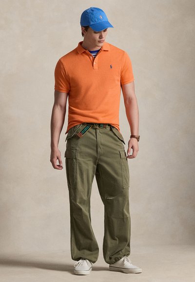 Νέος άνδρας που φοράει πορτοκαλί πόλο, μπλε καπέλο, ριγέ πουκάμισο από μέσα, χακί παντελόνι cargo, λευκά αθλητικά και ρολόι χειρός, όρθιος με τα χέρια χαλαρά.