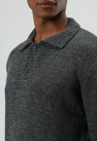 Grijze gebreide trui met een omgevouwen kraag, geribbelde structuur en vier zwarte knopen langs de knoopsluiting. Gladde stof met een subtiele glans.