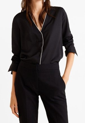 Femme portant un chemisier noir à manches longues avec passepoil blanc et un pantalon noir taille haute, debout avec une main dans la poche.