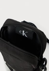 Fekete nylon keresztpántos táska cipzáras nyílással. A belsejében logó címke található. Pánttal és fémes díszítő elemekkel ellátva.