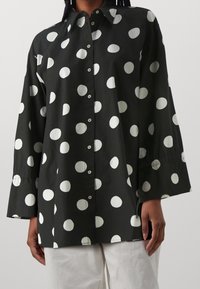 Chemise noire à grands pois blancs, dotée d'un col pointu, de manches longues et d'une fermeture boutonnée sur le devant, fabriquée en tissu léger.