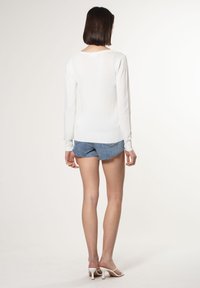 Weißes Langarmoberteil aus leichtem Strickmaterial, runder Halsausschnitt, rippenbesetzte Bündchen, getragen mit Jeansshorts und weißen Absatzsandalen.