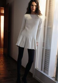 Abito bianco a costine con orlo svasato, maniche lunghe, abbinato a leggings neri e tacchi. Tessuto morbido, design minimalista.