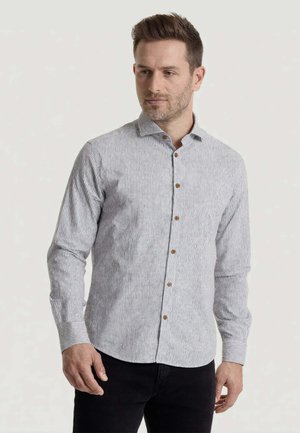 Uomo che indossa una camicia a righe grigio chiaro e bianca con bottoni marroni e pantaloni neri, in piedi davanti a uno sfondo semplice.