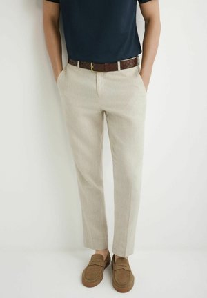 Mężczyzna ubrany w beżowe spodnie, brązowy pleciony pasek, brązowe zamszowe mokasyny i ciemnoniebieską koszulę wpuszczoną w spodnie, stojący na jednolitym białym tle.