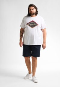 Witte katoenen t-shirt met een rood en zwart grafisch ontwerp, gecombineerd met donkerblauwe shorts en witte sneakers. Het model staat voor een neutrale achtergrond.