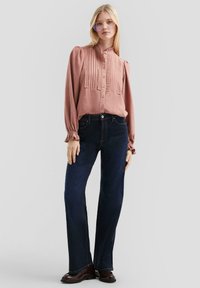 Blouse rose à manches longues avec des plis texturés et des boutons, associée à un jean bleu foncé évasé, portée avec des chaussures foncées. Fond gris clair.
