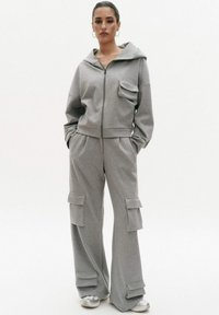 Szary zestaw dresowy z kapturem i spodniami cargo. Hoodie ma zamek błyskawiczny z przodu i jedną kieszeń na piersi. Spodnie mają duże kieszenie boczne i luźny krój.