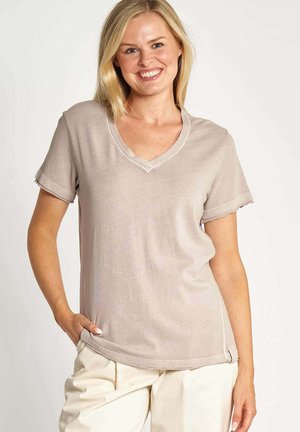 Blonde woman smiling, wearing a light beige short-sleeve V-neck shirt and off-white pants, with one hand in pocket against plain background.