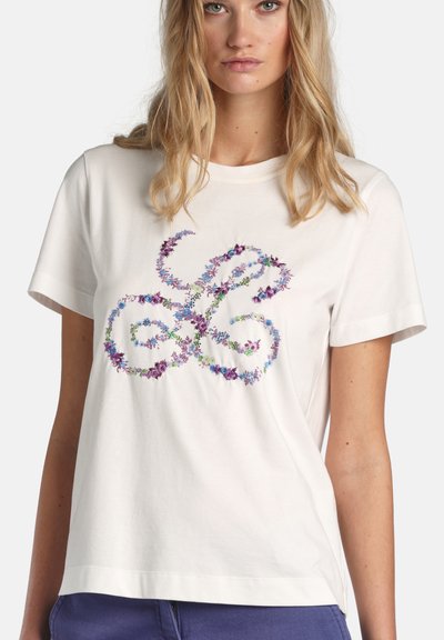 Nő, aki fehér, rövid ujjú inget visel, amelyen lila és kék virágminták absztrakt, hurkos mintázatot alkotnak az elején.