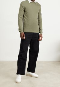 Un pull à manches longues vert olive avec un col rond, associé à un pantalon noir à jambes droites et des baskets blanches, présenté sur un fond neutre.