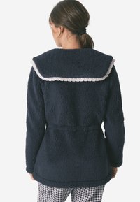 Cappotto in pile blu navy con un ampio colletto rotondo impreziosito da un bordo in pizzo bianco. Il cappotto ha una vita con cintura e un tessuto morbido e testurizzato.