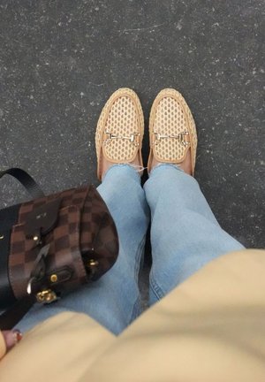 Voeten met beige gevlochten loafers en lichtblauwe spijkerbroek staand op donker asfalt, met een bruine geruite handtas zichtbaar.