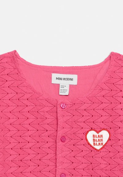 Haut en tricot rose avec motif zigzag, trois boutons à l'avant et un patch en forme de cœur portant l'inscription « BLAH BLAH BLAH » sur le côté droit de la poitrine.